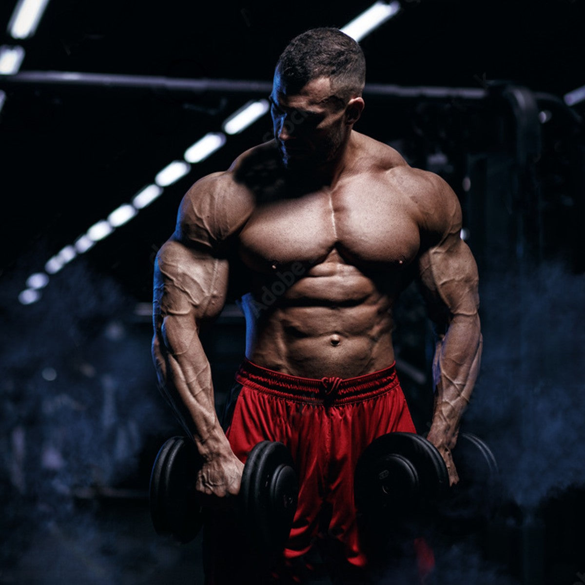 a lean and muscular male bodybuilder holding dumbbells by his side wearing red shorts.