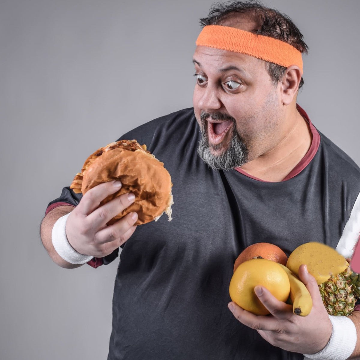 overweight man smiling at a burger while holding fruit in his arm.