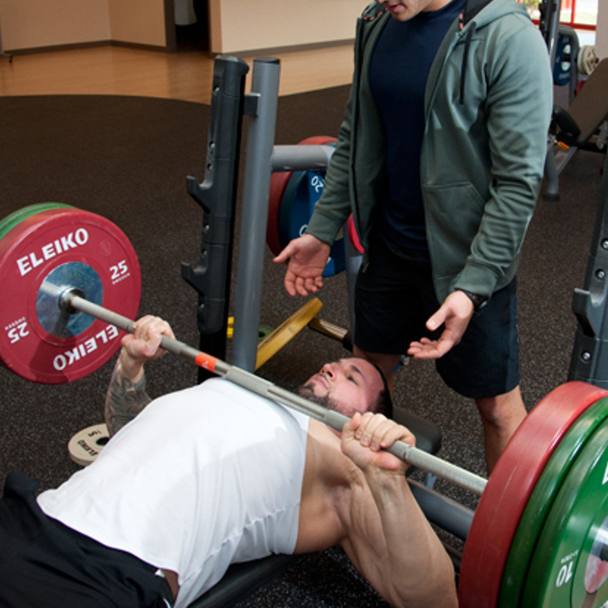 a male gym member performing a barbell bench press with a spotter behind him.