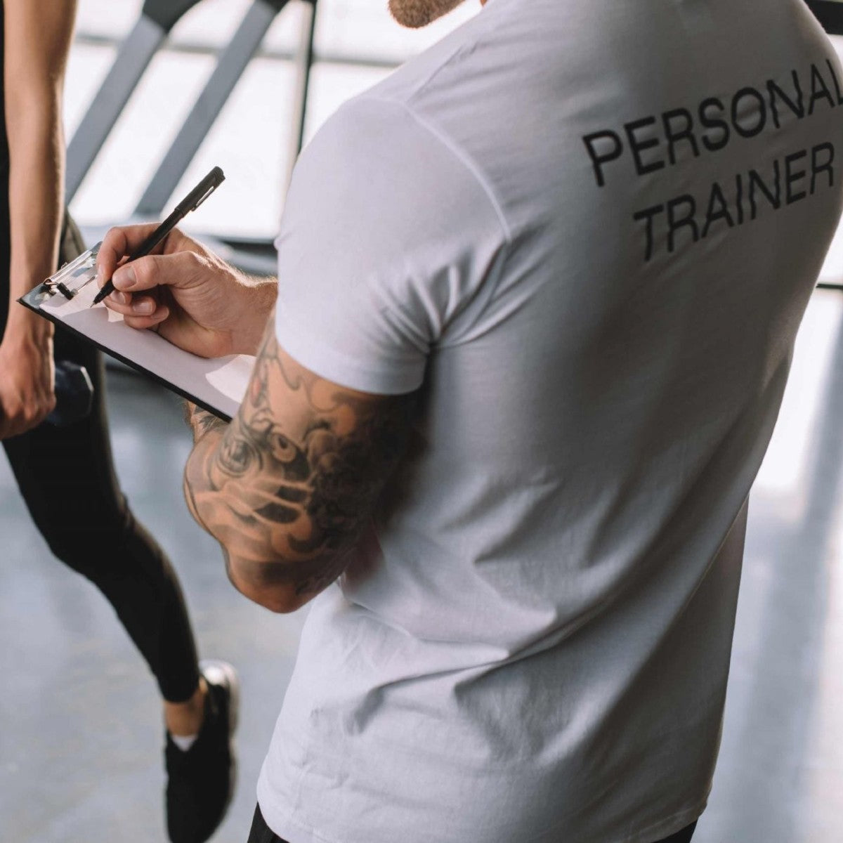 a personal trainer in a gym coaching a client wearing a white t-shirt with the words personal trainer on his back.